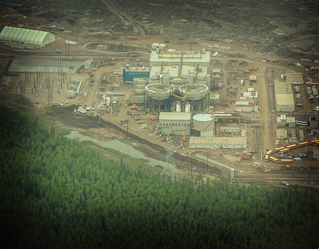 A bird's-eye view of a Fort McMurray industrial plant, next to a dense boreal forest.